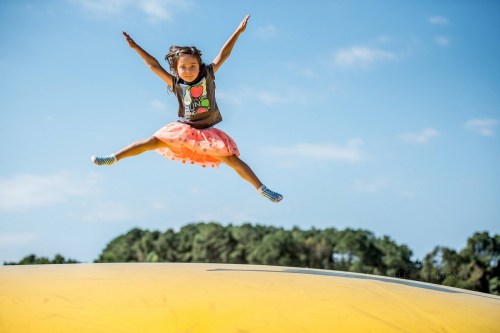 A young girl in a colorful outfit jumps joyfully in the air on a large inflatable surface against a blue sky.