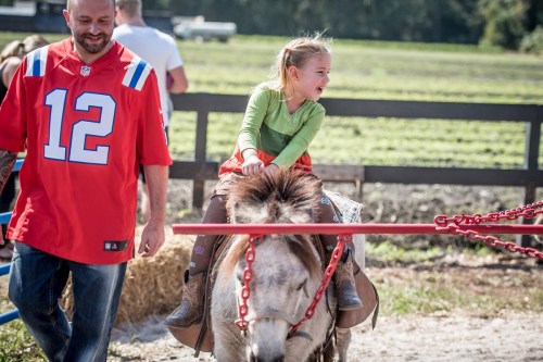 A girl in a green shirt joyfully rides a pony, while a man in a red jersey walks beside her on a sunny day.