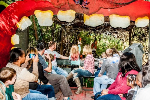 A group of people on a ride, seated inside a large, colorful mouth structure, surrounded by greenery.