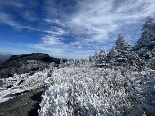Snow-covered trees and rocky terrain under a blue sky with wispy clouds, showcasing a winter landscape.