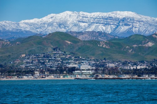 A coastal view featuring a snowy mountain range in the background and a green hillside with a small town by the water.
