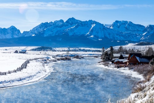 A serene winter landscape featuring a river, snow-covered mountains, and a cozy cabin under a clear blue sky.