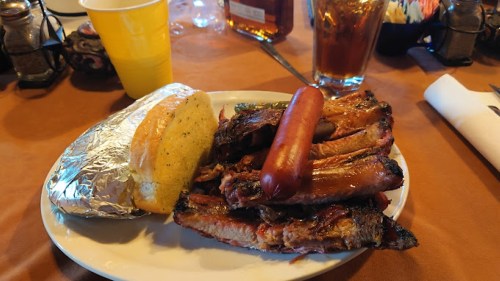 A plate of grilled ribs, a hot dog, and a foil-wrapped baked potato, with drinks in the background.
