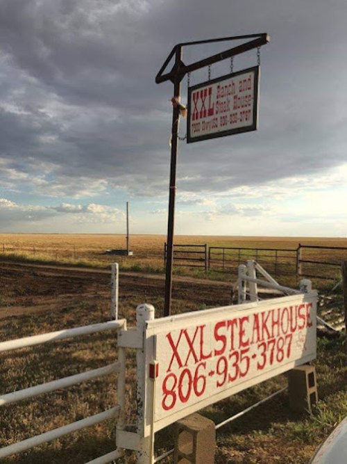 Sign for XXL Steakhouse and Ranch under a cloudy sky, with open fields in the background.