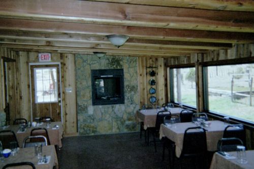 Cozy restaurant interior with wooden walls, tables set for dining, and a stone fireplace. Large windows provide natural light.
