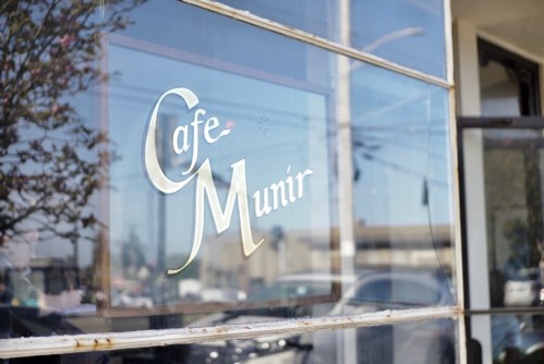Sign for "Cafe Munir" reflected in a window, with a blurred street scene in the background.