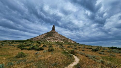 A tall rock formation rises from a grassy landscape under a cloudy sky, with a winding path leading towards it.