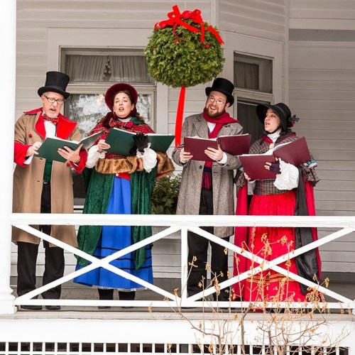 A group of four carolers in festive attire sing on a porch, with a large mistletoe hanging above them.