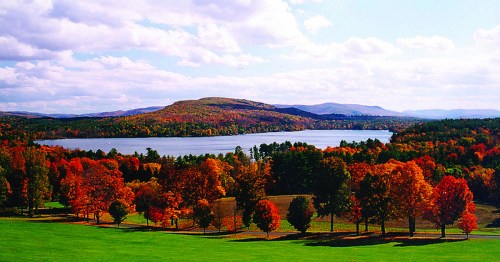 A scenic view of a lake surrounded by vibrant autumn foliage and rolling hills under a partly cloudy sky.