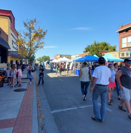 A bustling street fair with vendors and visitors, colorful tents lining the road under a clear blue sky.