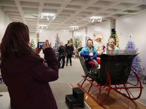 A woman takes a photo of two girls sitting in a sleigh with Santa in a festive room decorated with Christmas trees.