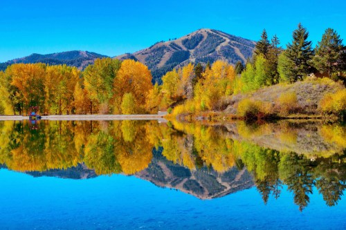 A serene lake reflecting vibrant autumn trees and a mountain under a clear blue sky.