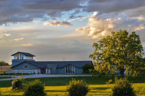 A modern house with large windows, surrounded by green grass and trees, under a cloudy sky at sunset.