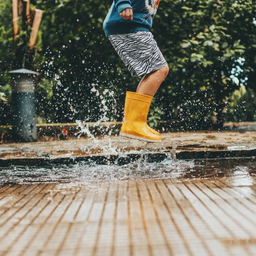 A child in a blue sweater and yellow rain boots splashes in a puddle on a rainy day.