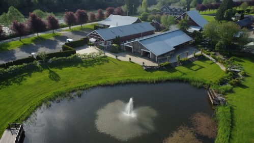 Aerial view of a serene property with a pond, fountain, and landscaped gardens surrounded by buildings and trees.