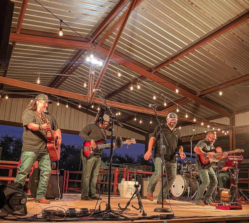 A band performs on stage under string lights, with musicians playing guitars and drums in a lively outdoor setting.
