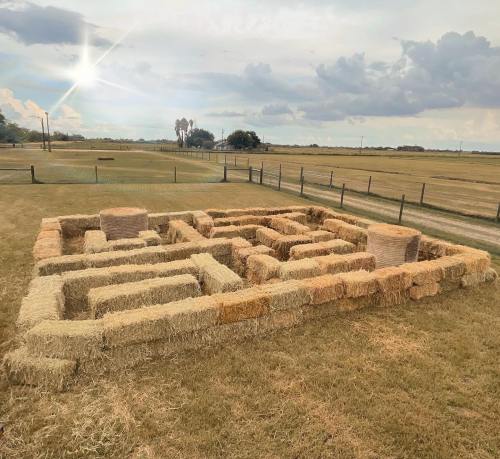A maze made of hay bales set in a grassy field under a cloudy sky with a hint of sunlight.