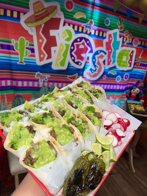 A colorful platter of tacos with guacamole, radishes, lime, and grilled peppers, set against a festive "Fiesta" backdrop.