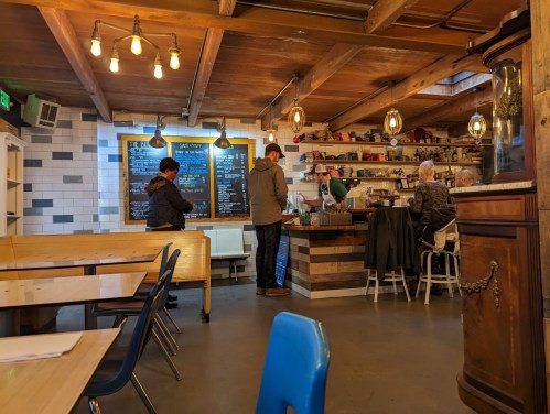 A cozy café interior with wooden beams, patrons at the counter, and a chalkboard menu on the wall.
