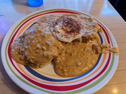 A colorful plate with biscuits and gravy topped with a fried egg, served on a wooden table.