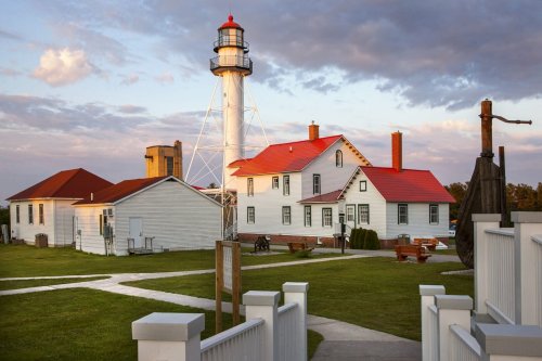 A lighthouse with a red roof stands beside a white building, surrounded by green grass and a cloudy sky.