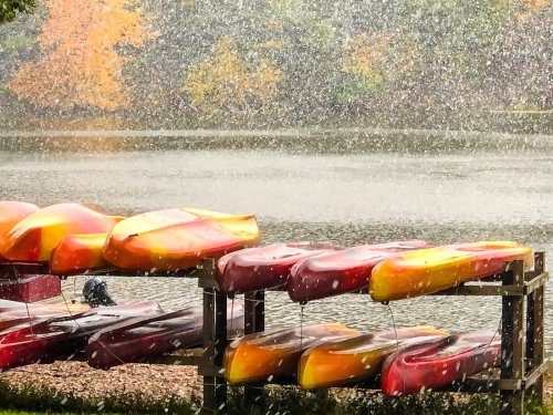Colorful kayaks stacked on a rack by a lake, with snowflakes falling and autumn trees in the background.