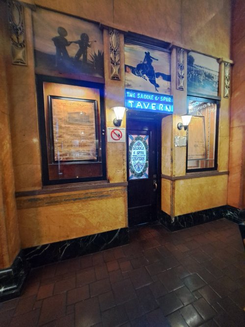 Entrance to a vintage tavern with ornate decor, featuring neon signs and framed artwork on the walls.