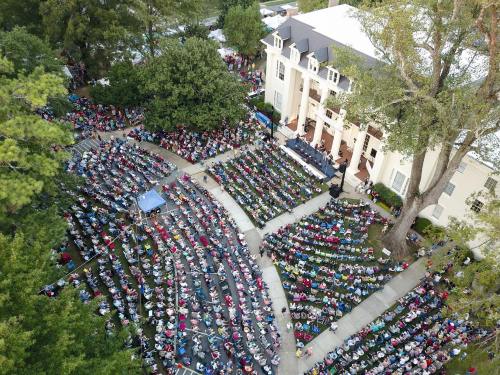 Aerial view of a large outdoor crowd seated in rows, gathered in front of a building for an event surrounded by trees.