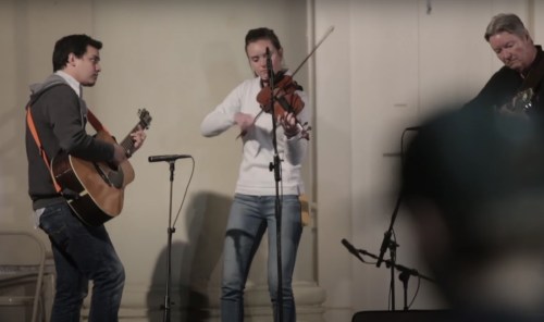 Three musicians perform together: one on guitar, one playing violin, and another on guitar, in a cozy setting.