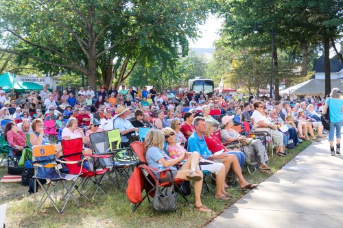 A large crowd of people seated in folding chairs at an outdoor event, surrounded by trees and tents.