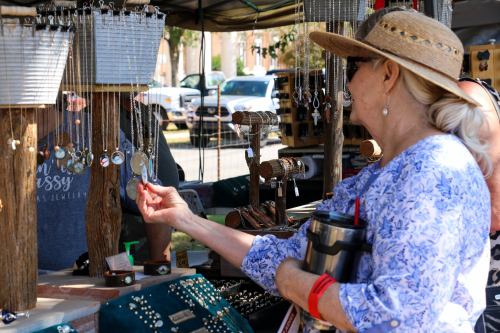 A woman in a straw hat examines jewelry at a market stall, holding a drink and surrounded by various necklaces.