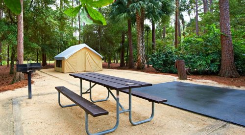 A campsite featuring a tent, picnic table, grill, and a paved area surrounded by trees and greenery.