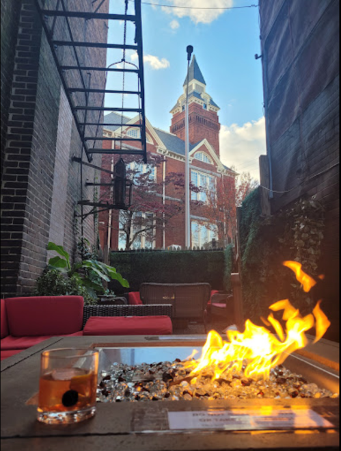 A cozy outdoor space with a fire pit, a cocktail, and a historic building in the background under a blue sky.