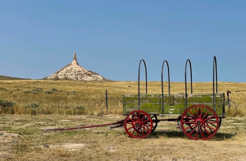 A vintage green wagon with red wheels sits in a grassy field, with a tall rock formation in the background under a clear blue sky.
