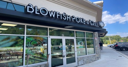 Exterior of Blowfish Poke & Grill restaurant, featuring large signage and glass doors, with a clear blue sky above.