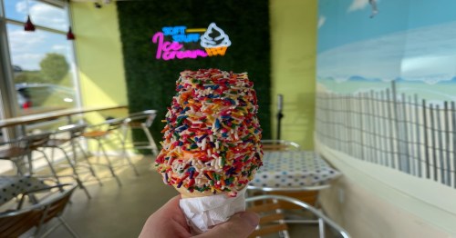 A hand holding a colorful ice cream cone covered in sprinkles, with a bright ice cream shop background.