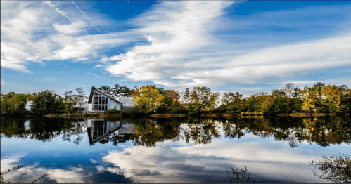 A serene lake reflects a modern building and colorful trees under a blue sky with wispy clouds.