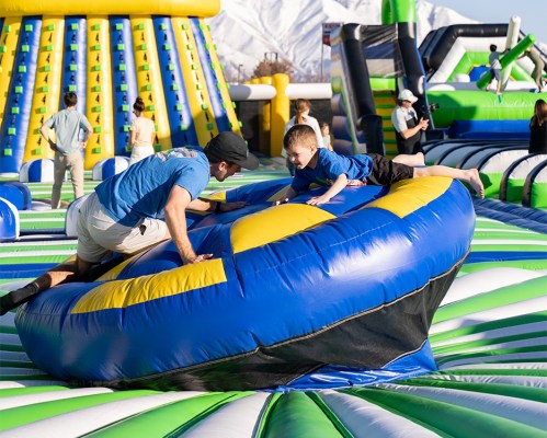A man and a child play on a large inflatable bounce pad at a fun outdoor event with colorful structures in the background.