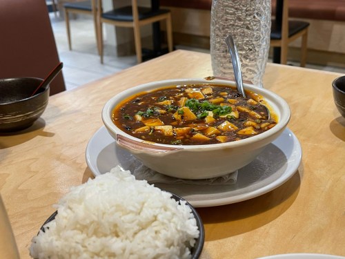 A bowl of mapo tofu with green onions, served with a side of white rice on a wooden table.