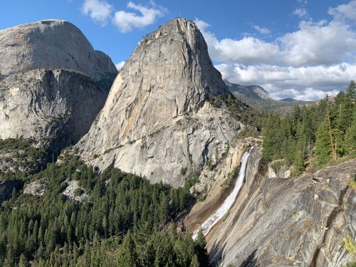A scenic view of towering granite cliffs and a waterfall surrounded by lush green forests under a blue sky.