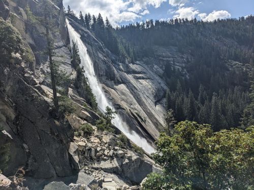 A waterfall cascades down a rocky cliff surrounded by lush green trees and mountains under a partly cloudy sky.