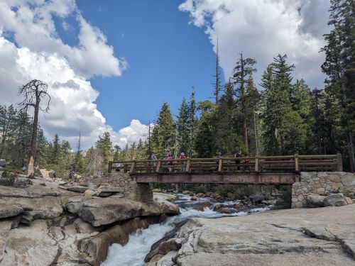 A wooden bridge spans a rocky river, surrounded by tall trees and a blue sky with fluffy clouds.