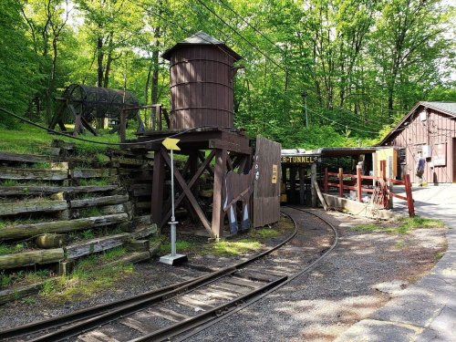 A wooden water tower beside a train track, surrounded by greenery and rustic buildings in a forested area.