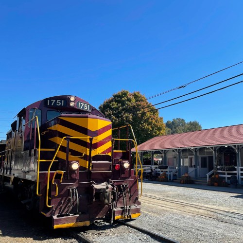 A vintage train engine parked near a station, surrounded by trees and a clear blue sky.