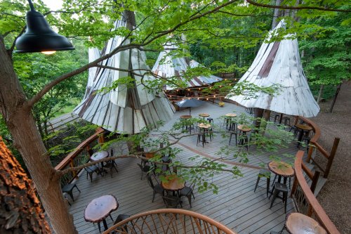 A wooden deck with tables surrounded by teepee-like structures and lush green trees.