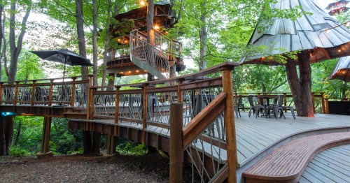 A wooden deck with a treehouse and dining area, surrounded by lush greenery and soft lighting.