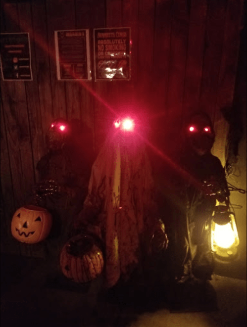 Three spooky figures with glowing red eyes, holding lanterns and pumpkins, set against a dark, wooden background.