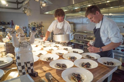 Two chefs plating dishes with mussels in a busy kitchen, with a ceramic chef figure in the foreground.