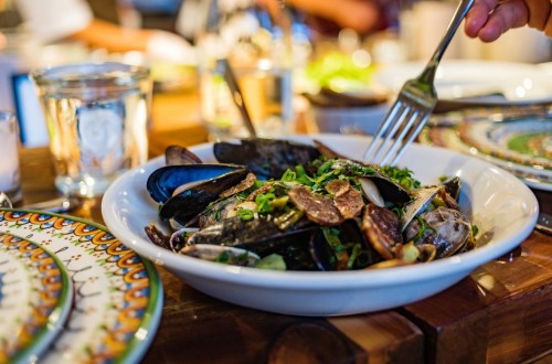 A bowl of mussels topped with herbs and spices, with a fork poised to serve, set on a wooden table.