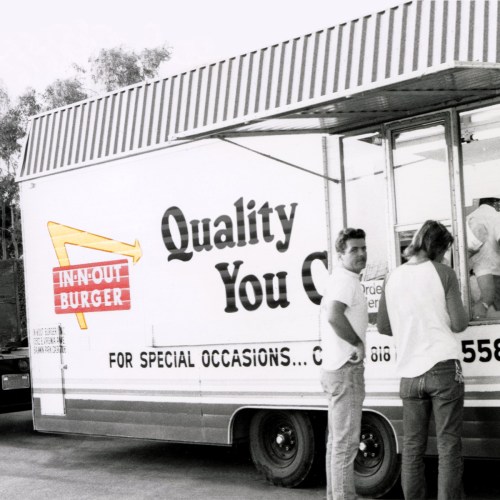 A vintage In-N-Out Burger food truck with two people standing outside, showcasing the logo and menu details.